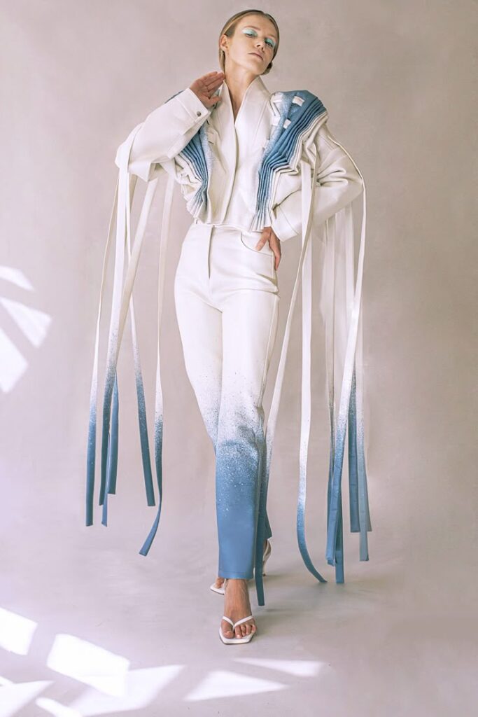 Fashion model in white outfit with blue ombré pants and flowing ribbons, striking modern look in studio.