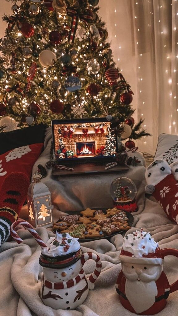 Cozy Christmas setup with tree, decorated cookies, hot chocolate in snowman and Santa mugs, and virtual fireplace.