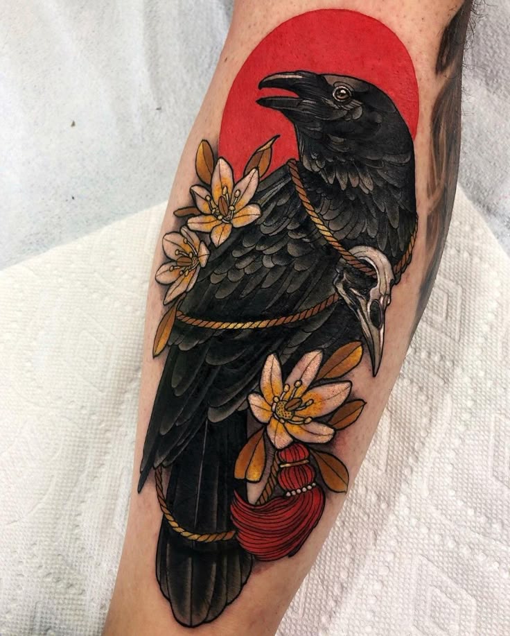 Raven tattoo design with flowers and red sun on arm, featuring a decorative skull and rope elements.