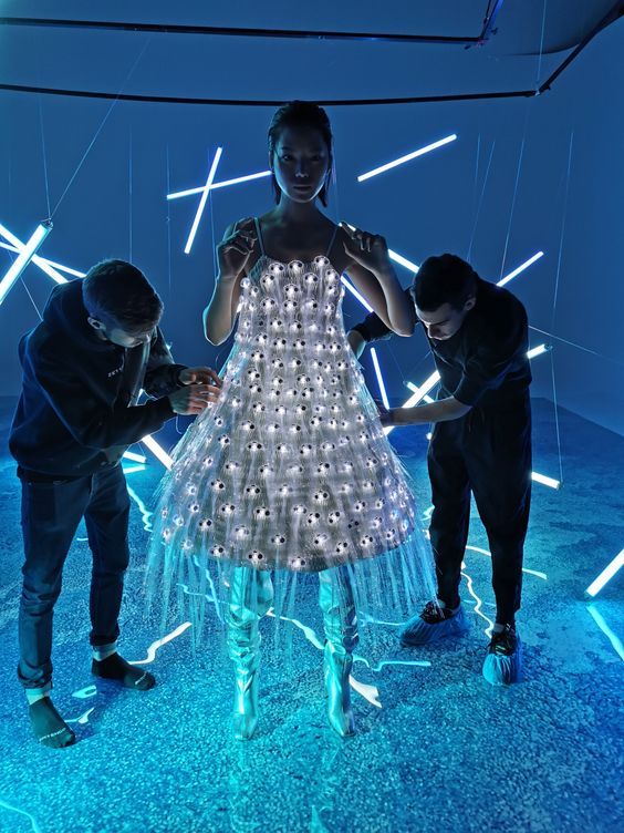 Model in LED light-up dress, futuristic setting, assisted by designers under neon blue lights.