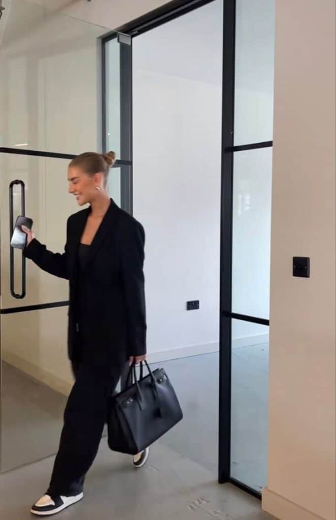 Person in black suit and sneakers entering modern office, holding a smartphone and handbag.