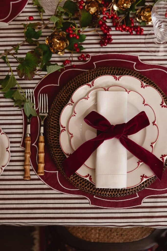 Festive table setting with elegant white plates, burgundy napkin, and holiday greenery on striped tablecloth.