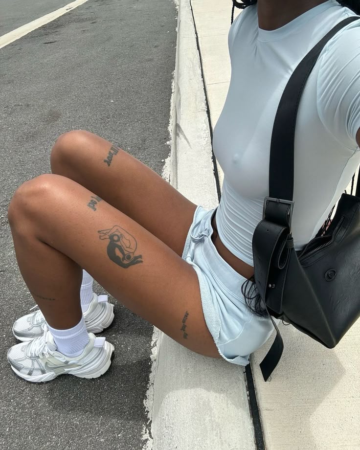 _ 16 Person sitting on a curb wearing a light blue outfit and white sneakers, showcasing leg tattoos and a black bag. | Sky Rye Design Person sitting on a curb wearing a light blue outfit and white sneakers, showcasing leg tattoos and a black bag.