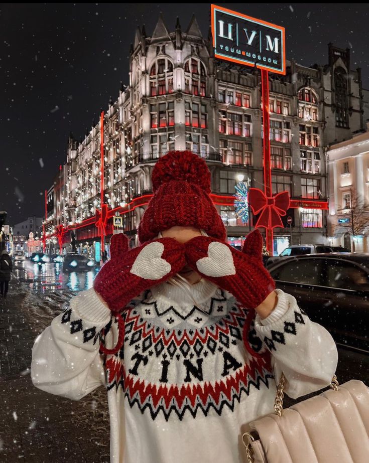 163b3279-89d4-48b4-8b8d-901b0efa82f0 Person in cozy winter attire making heart shape with mittens in snowy city street near decorated building at night. | Sky Rye Design Person in cozy winter attire making heart shape with mittens in snowy city street near decorated building at night.