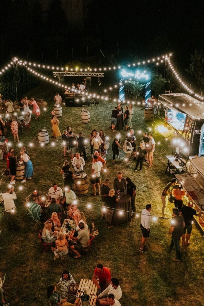 Outdoor night market event with string lights and people socializing, enjoying food and drinks from food trucks.