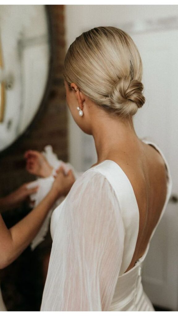 Bride with elegant bun hairstyle and pearl earrings in a satin dress, preparing for wedding ceremony.