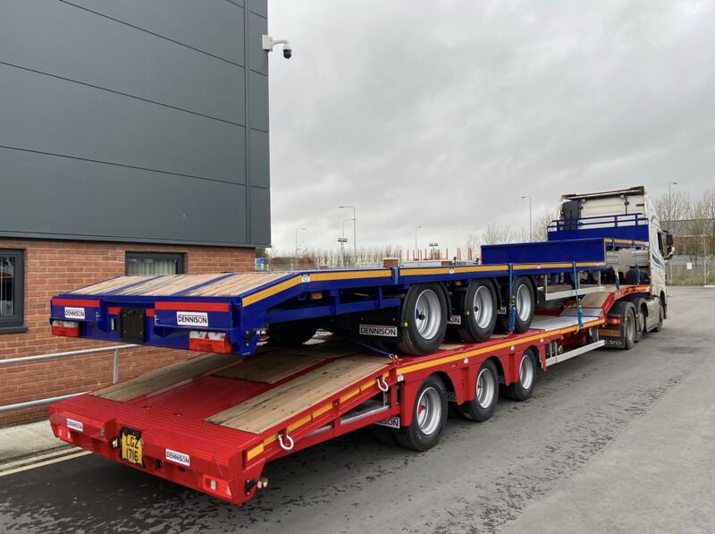 Truck transporting blue and red flatbed trailers, parked near a building on a cloudy day.