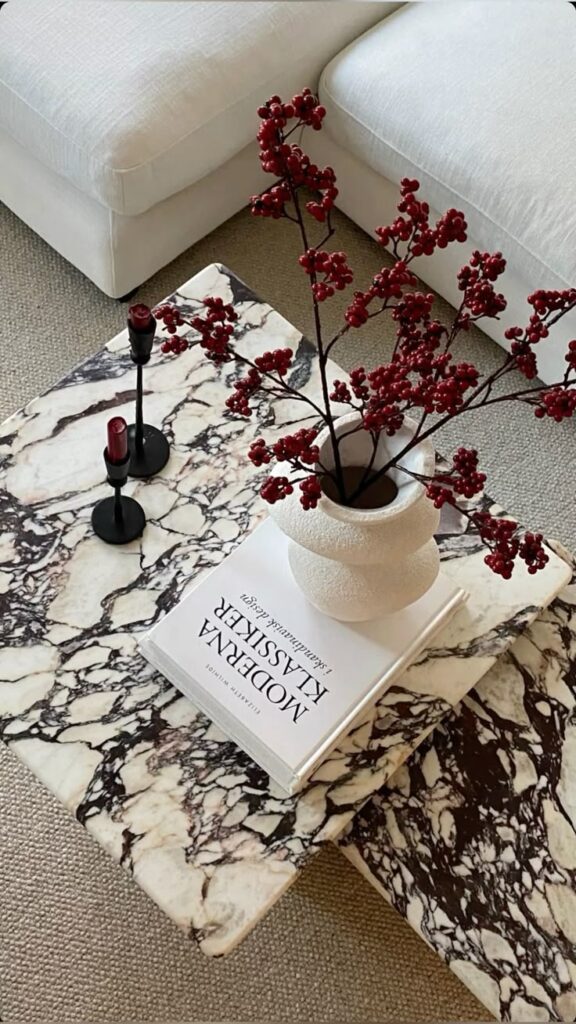 178807bc-f3ab-45d8-b703-076f68bad62c Marble coffee table with red berry vase, book, and candles, next to a white sofa. Stylish modern home decor setting. | Sky Rye Design Marble coffee table with red berry vase, book, and candles, next to a white sofa. Stylish modern home decor setting.