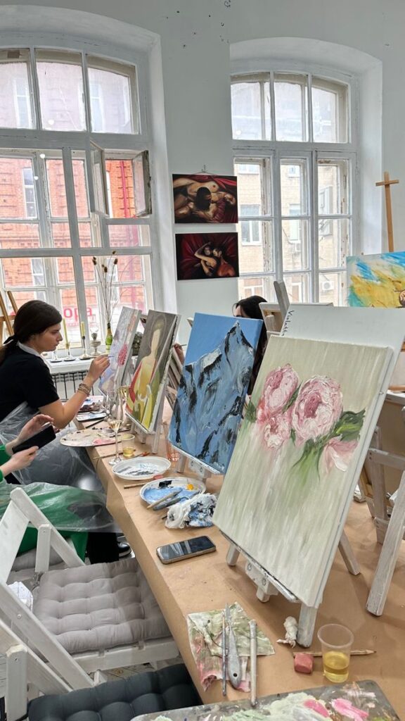Artists painting in a bright studio with canvases of flowers and mountains on easels.