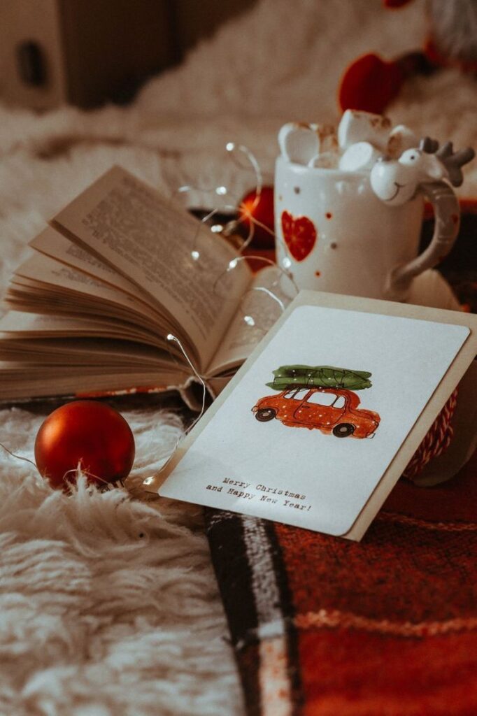 _ 18 Cozy Christmas scene with an open book, festive mug, card, and red ornament on a blanket. | Sky Rye Design Cozy Christmas scene with an open book, festive mug, card, and red ornament on a blanket.