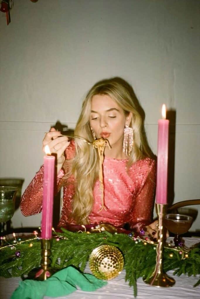 Woman in pink dress enjoying pasta at a candlelit dinner table, with festive decorations and long earrings.