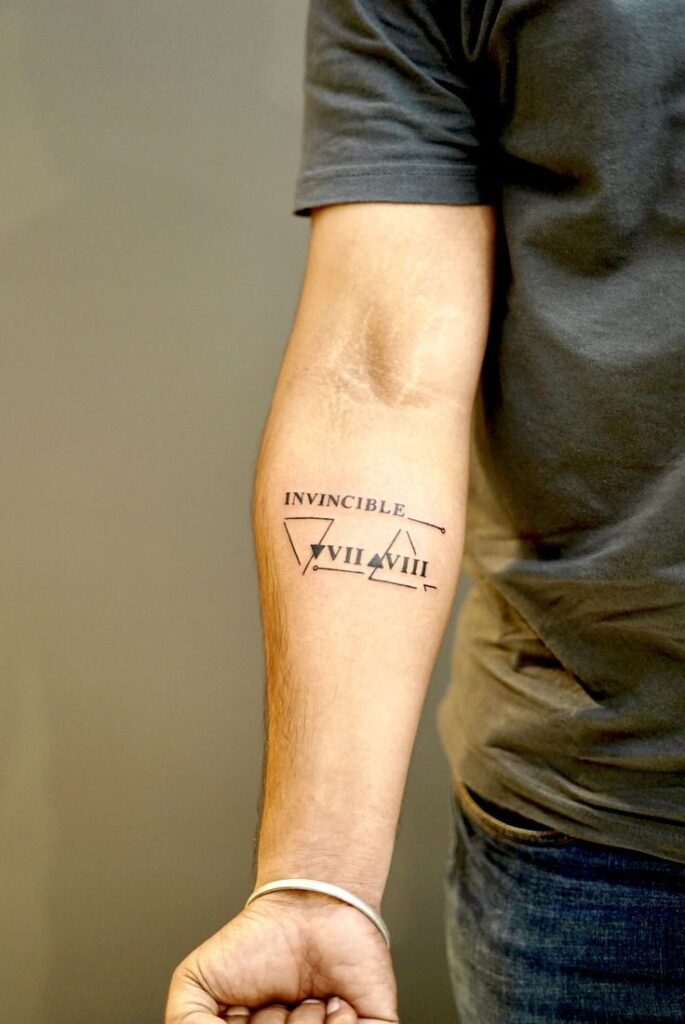 Forearm tattoo with Invincible text and Roman numerals, featuring geometric designs on a man's arm.