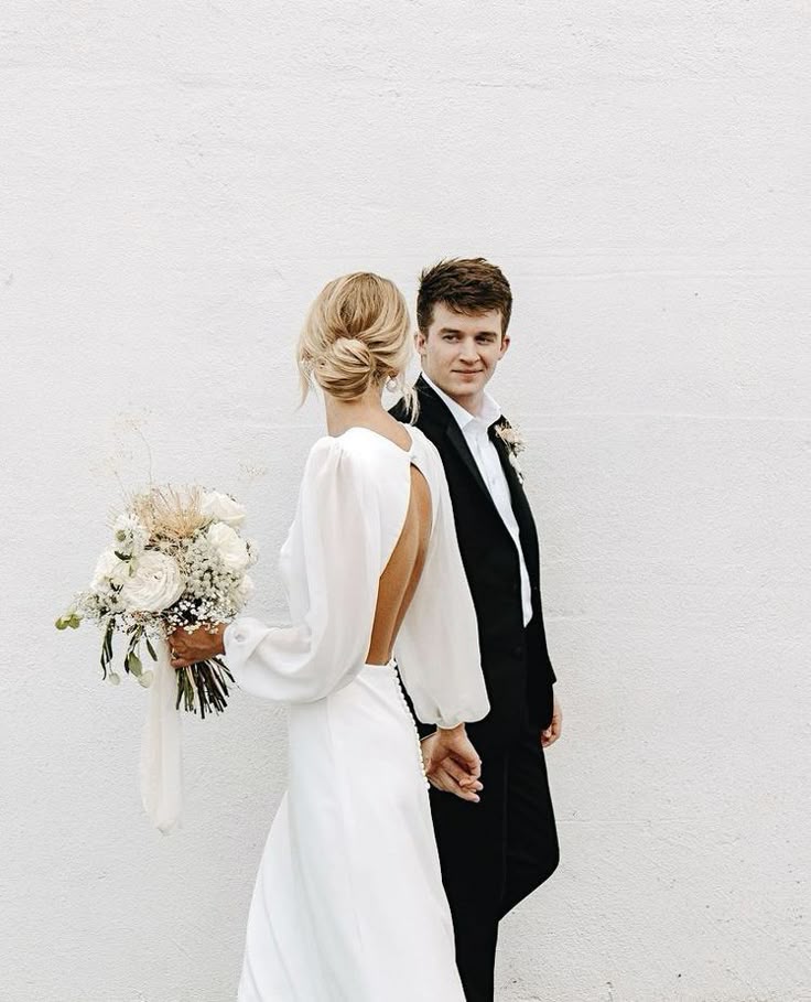 Bride and groom holding hands, dressed stylishly for their wedding day against a white backdrop.