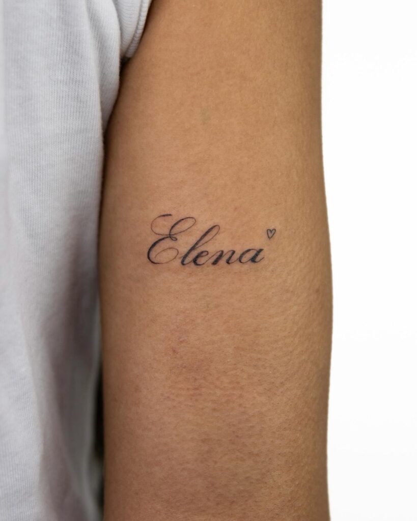 Arm tattoo with the name Elena in elegant script, featuring a small heart symbol. Minimalist black ink design.