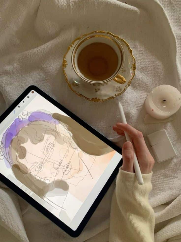 Digital art creation on tablet with stylus, tea cup, and candle on cozy blanket background.