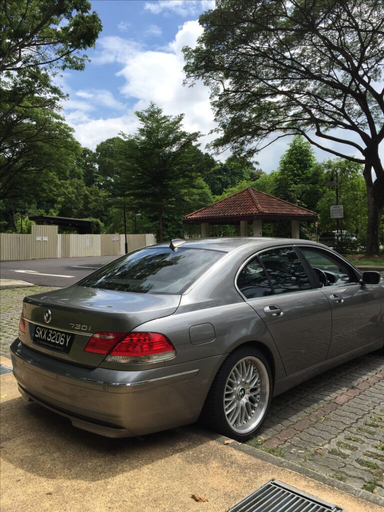 Silver BMW 730i parked outdoors, surrounded by lush greenery and a tiled roof structure in the background.