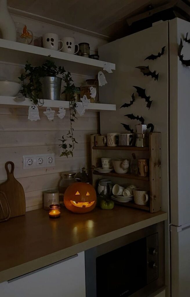 Cozy Halloween kitchen with jack-o'-lantern, ghost decor, bats, and mugs on wooden shelves. Warm candlelight glow.