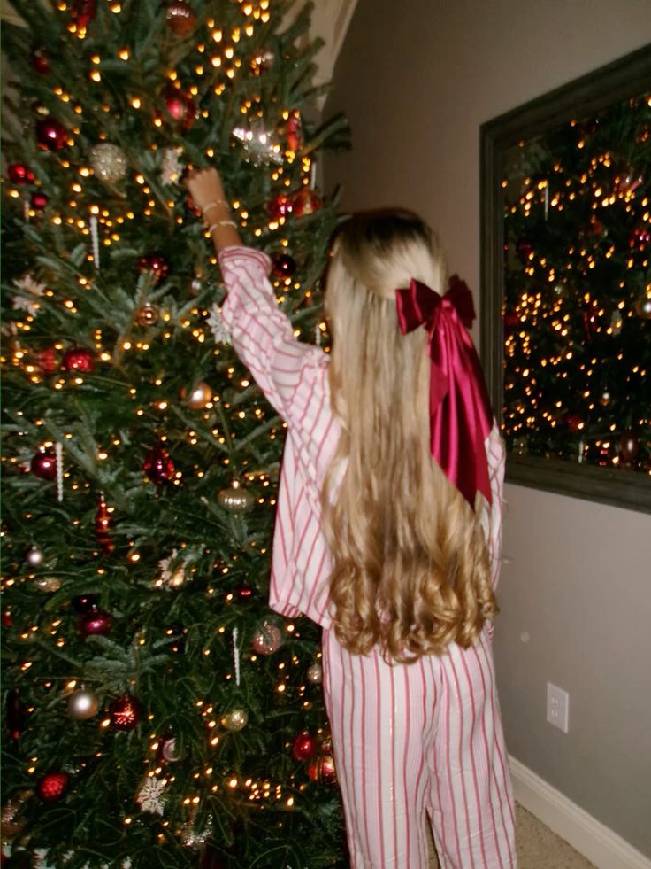 Woman in red-striped pajamas decorates a Christmas tree with ornaments, wearing a red bow in her hair.