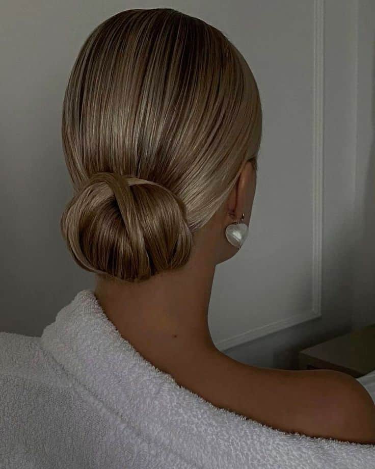 Elegant blonde hair styled in a sleek low bun, highlighted in soft lighting, with pearl earring detail.