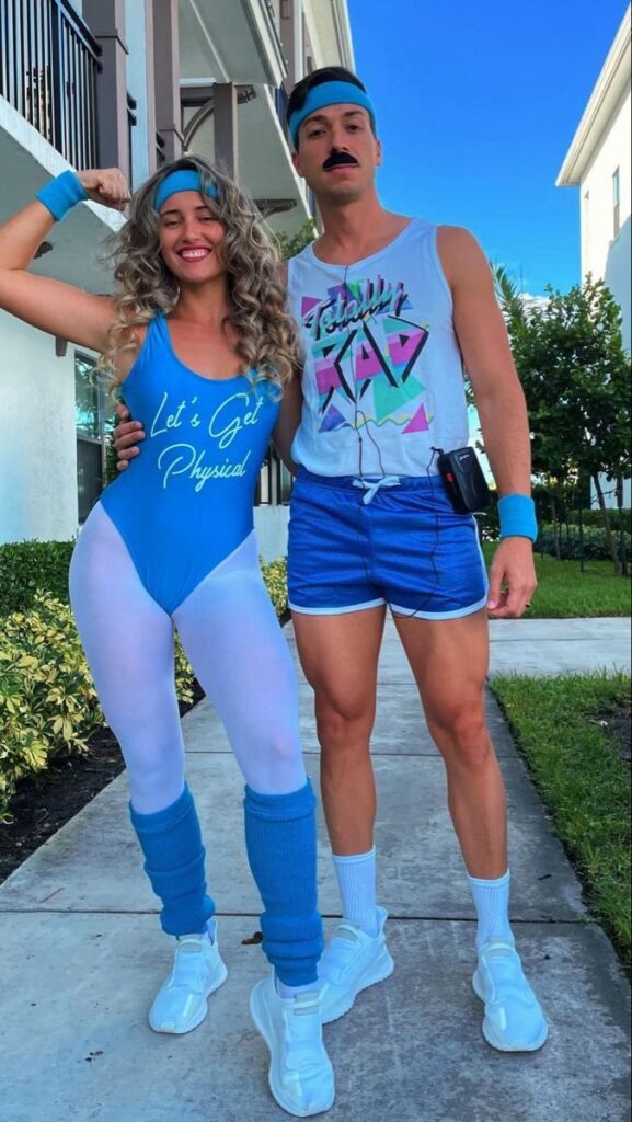 Couple in 80s workout costumes, smiling outdoors. Woman flexes, wearing blue leotard. Man in tank top with retro design.