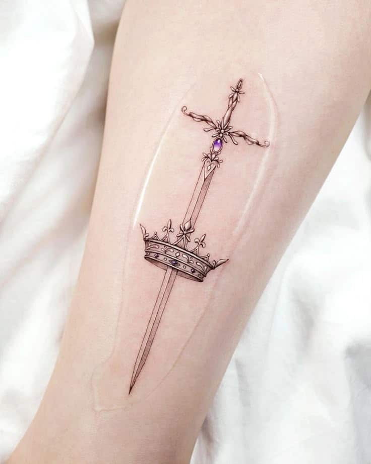 _ 2 Elegant sword and crown tattoo design on forearm with intricate details, featuring a purple gem accent. | Sky Rye Design Elegant sword and crown tattoo design on forearm with intricate details, featuring a purple gem accent.