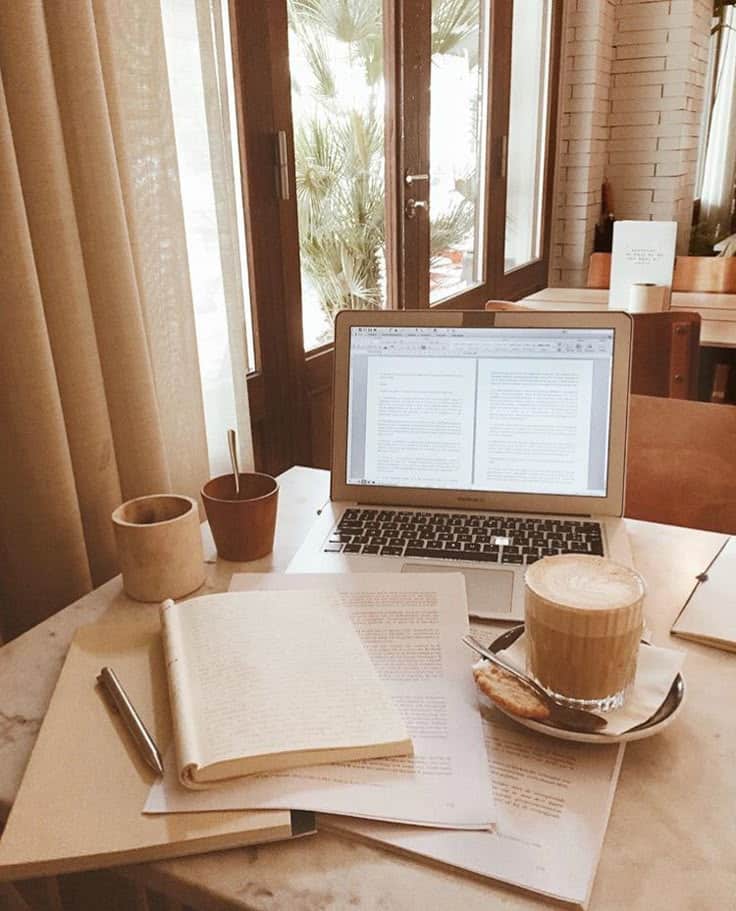 Cozy workspace with laptop, open book, and coffee cup on a marble table by the window in a warm, inviting cafe setting.