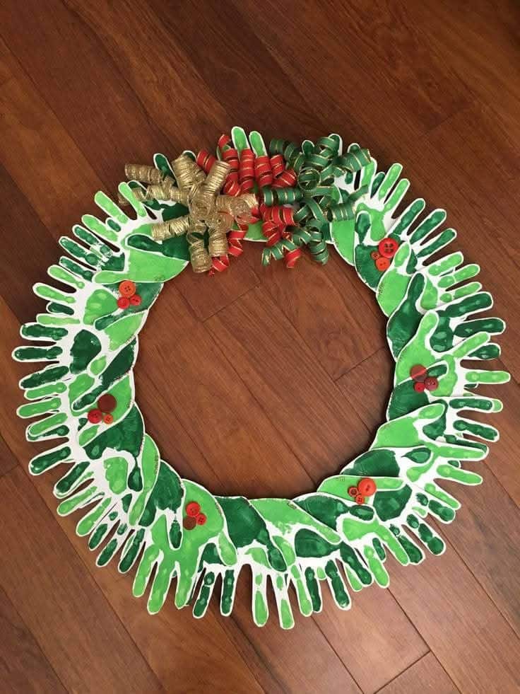 Handprint wreath with green hands, red buttons, and curly ribbons on a wooden floor, crafting holiday decoration.