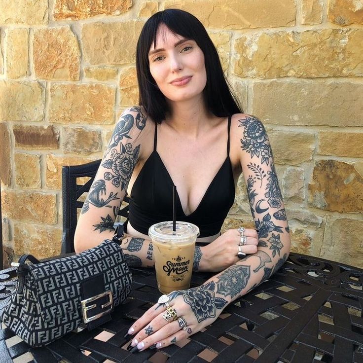 Woman with tattoos enjoying iced coffee outdoors, sitting at a table with purse, against stone wall backdrop.