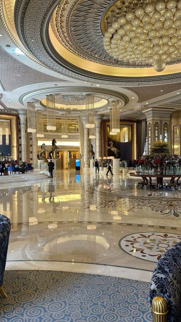 Luxurious hotel lobby with ornate ceiling, marble floors, elegant furnishings, and grand chandelier.