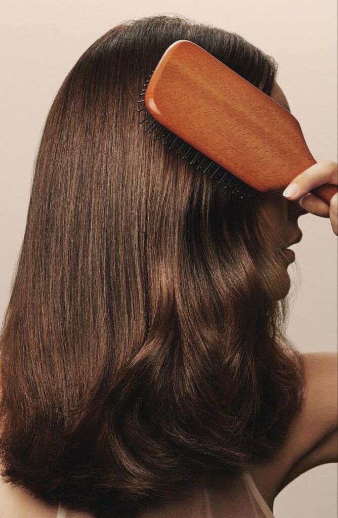 Woman brushing long, smooth brown hair with wooden brush for hair care.