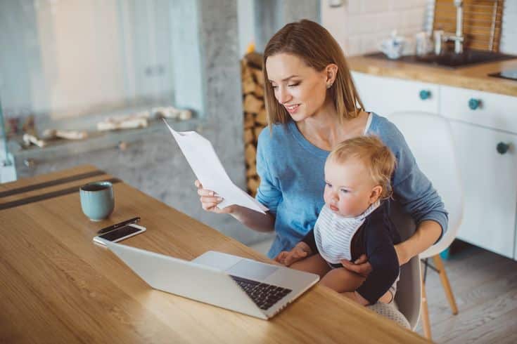 Mother multitasking with baby, reviewing documents, and working on laptop in kitchen.