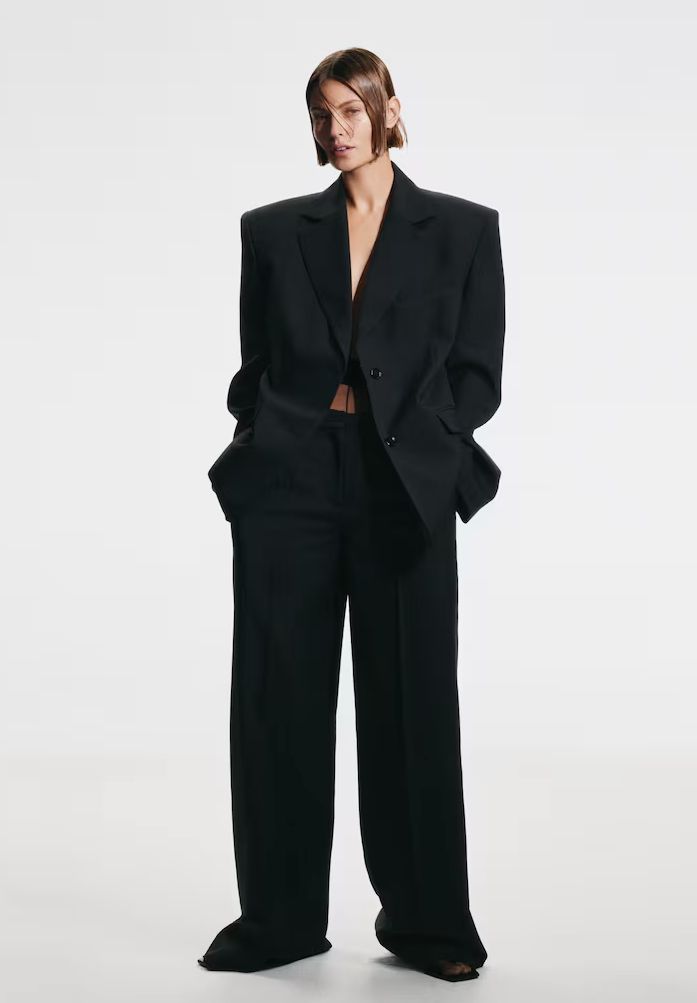 Fashion model in oversized black suit, hands in pockets, against white background.