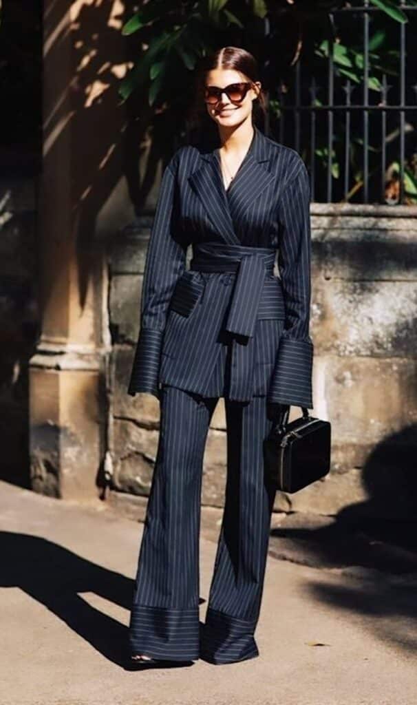 _ 20 Woman in a stylish pinstripe suit and sunglasses, holding a purse outdoors in bright sunlight. | Sky Rye Design Woman in a stylish pinstripe suit and sunglasses, holding a purse outdoors in bright sunlight.