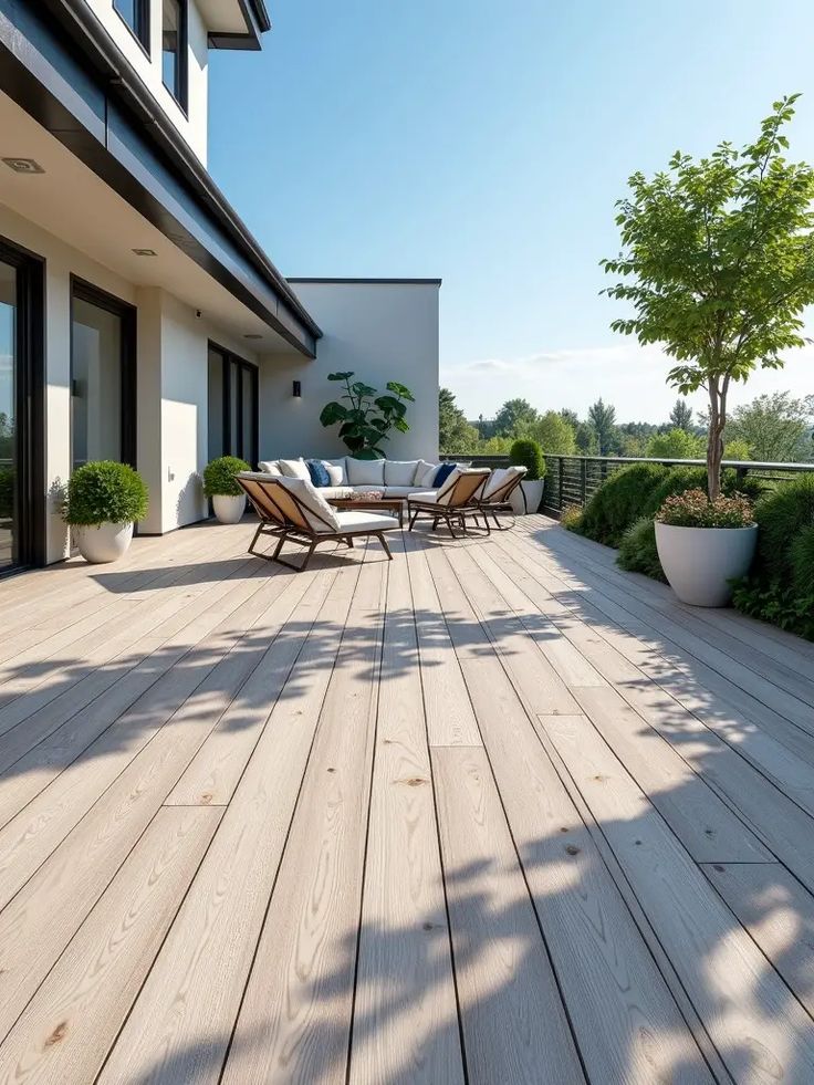 Modern outdoor patio with wooden decking, cozy chairs, and lush greenery under a clear blue sky.