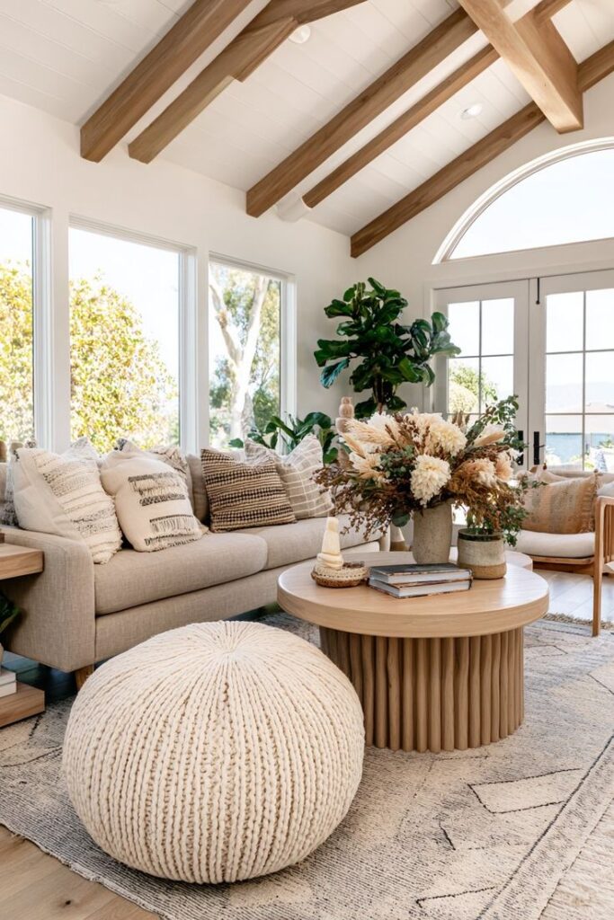 Cozy living room with beige sofa, knitted pouf, and wooden coffee table, featuring boho-style decor and large windows.