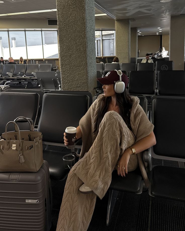 Woman in cozy knitwear with headphones, coffee, and luggage at the airport, seated and waiting at the gate.