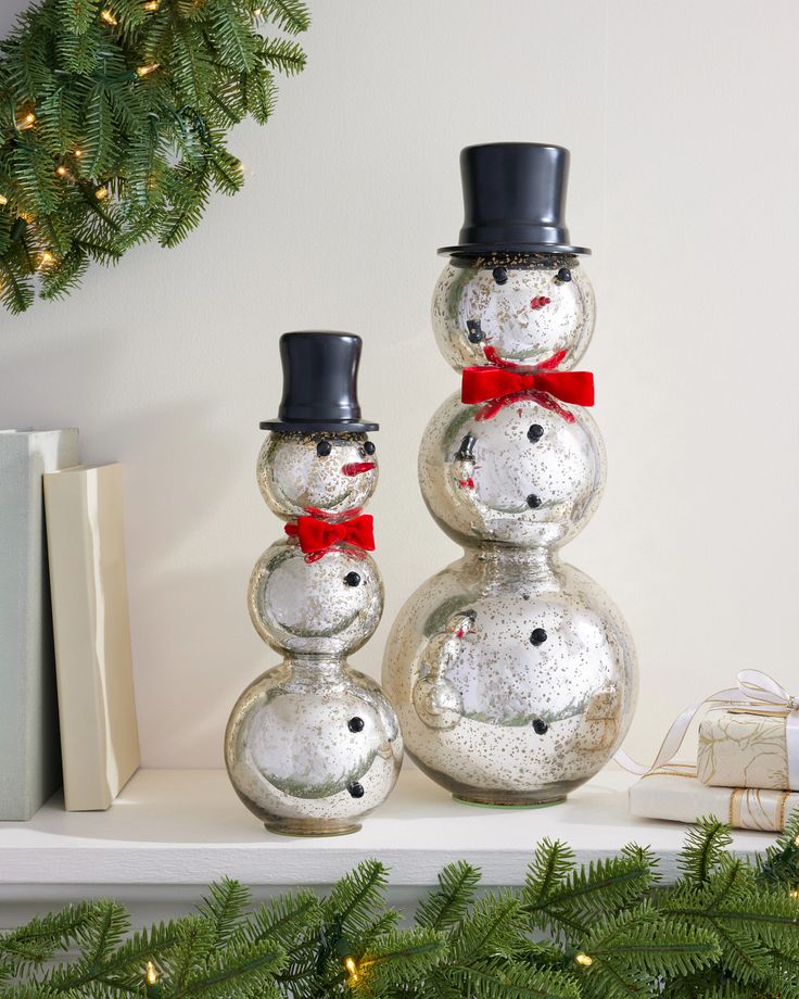 20_ Silver Tabletop Mercury Glass Snowmen Set of 2 Width 8 in by Balsam Hill Glass snowman figurines with black hats and red bows on a shelf, surrounded by festive greenery and a gift box. | Sky Rye Design Glass snowman figurines with black hats and red bows on a shelf, surrounded by festive greenery and a gift box.