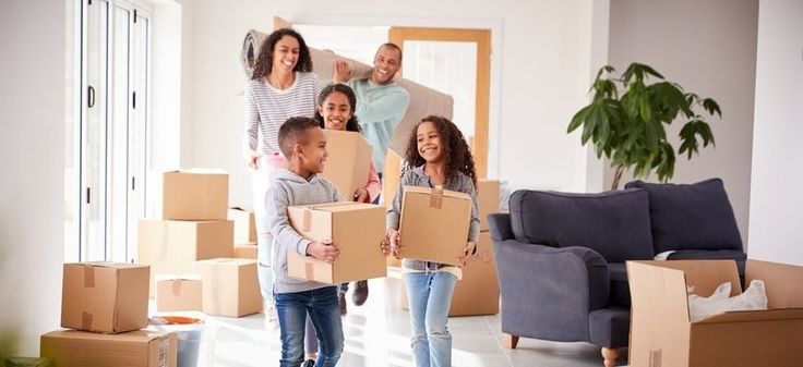 Family moving into a new home, carrying boxes and smiling, surrounded by unpacked cardboard boxes in the living room.