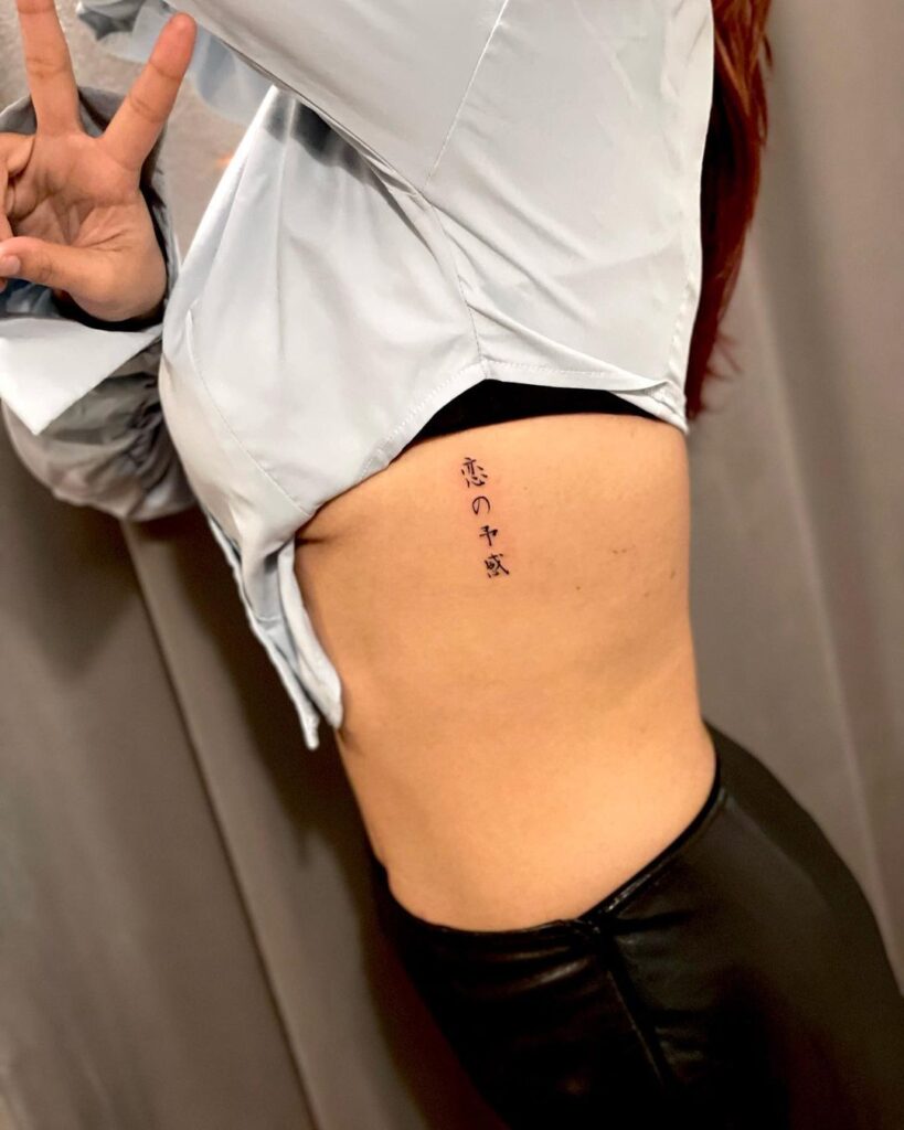 Side view of a person showing a Japanese tattoo on their ribs, wearing a white shirt and black pants, making a peace sign.