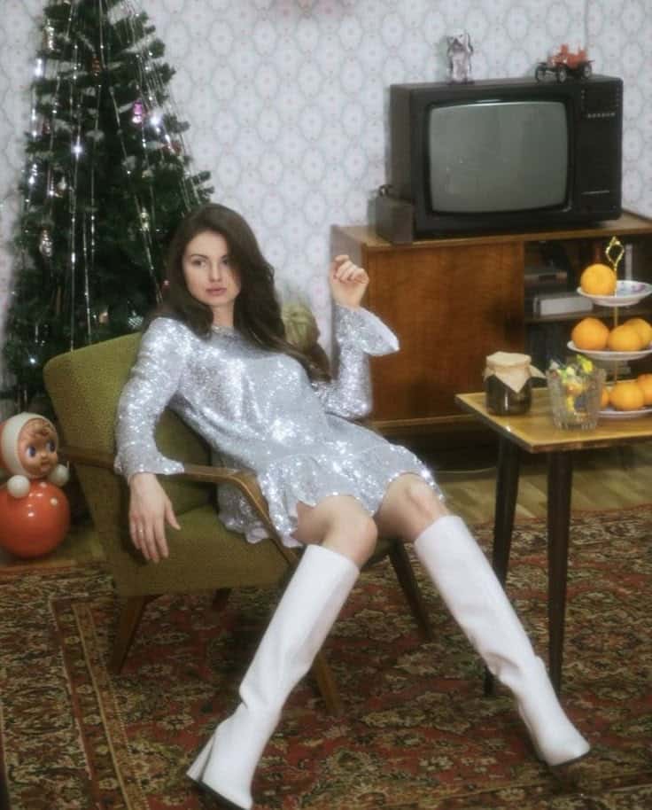 Woman in retro silver dress and boots, lounging by a vintage TV, Christmas tree, and festive decor. Retro holiday vibe.