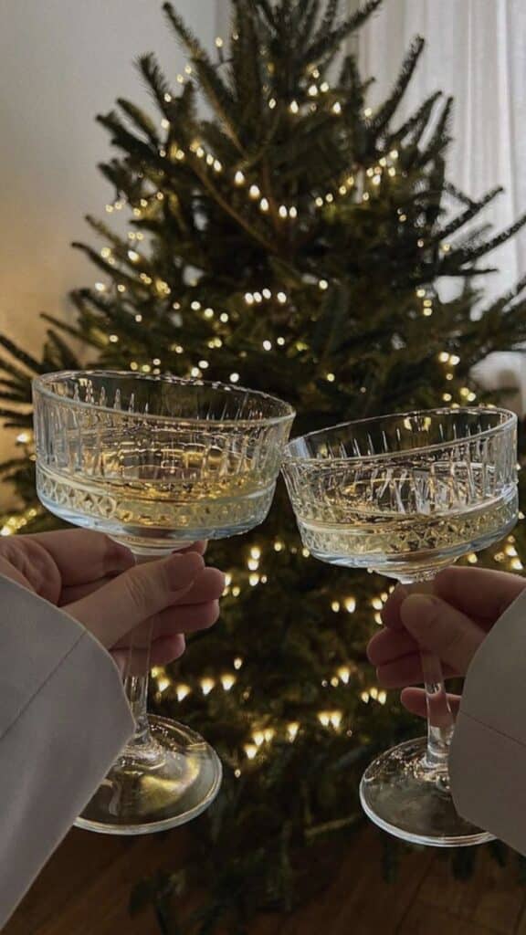 Screenshot Two hands clinking crystal glasses in front of a decorated Christmas tree with warm lights. | Sky Rye Design Two hands clinking crystal glasses in front of a decorated Christmas tree with warm lights.