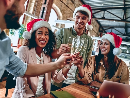 Colleagues wearing Santa hats celebrate with champagne at an office Christmas party, smiling and toasting.