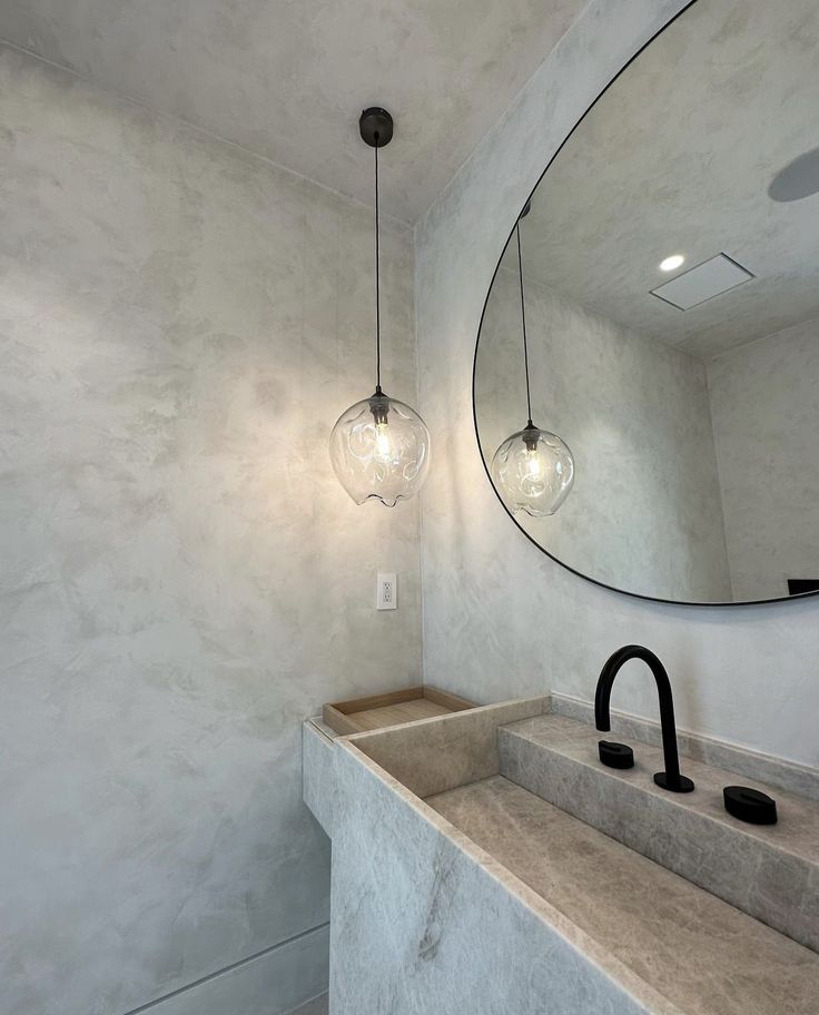 Minimalist bathroom with stone sink, black faucet, round mirror, and elegant pendant lighting.