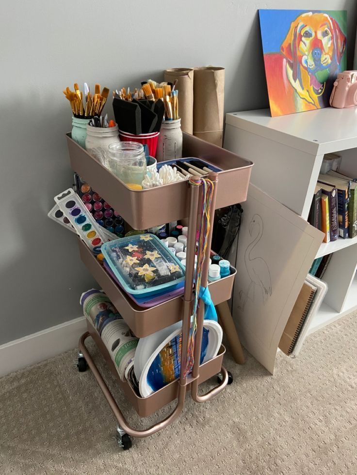 Art supplies organized on a tiered cart with brushes, paints, and canvas in a creative workspace.