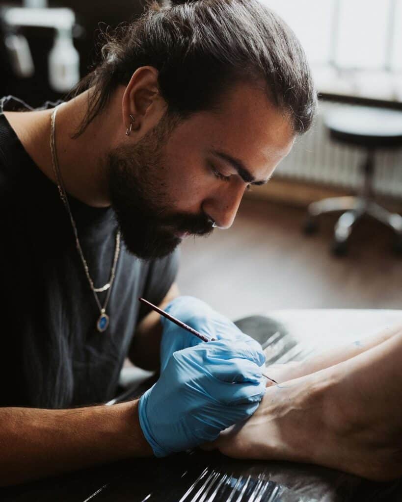 2379c80e5237997c3132108168affc71 Tattoo artist focused on inking a foot, wearing blue gloves in a well-lit studio. | Sky Rye Design Tattoo artist focused on inking a foot, wearing blue gloves in a well-lit studio.