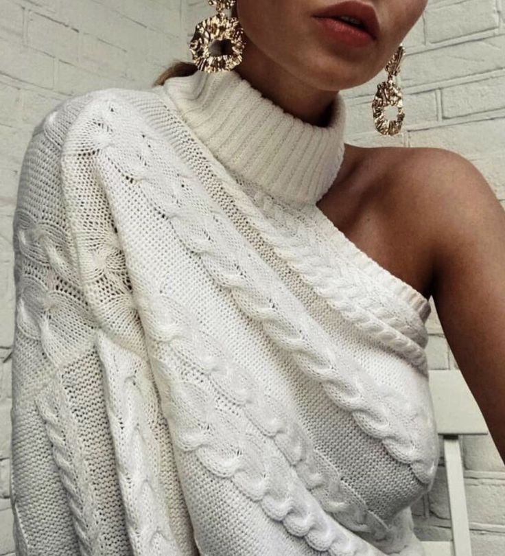 Stylish woman in white cable knit sweater and gold earrings against a brick backdrop.