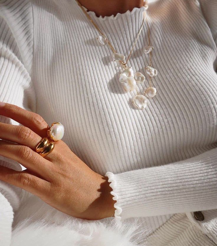 Woman wearing white ribbed sweater with pearl necklaces and gold rings, showcasing elegant jewelry style.