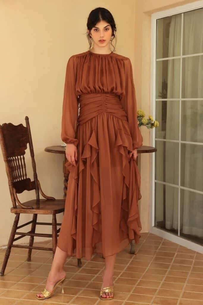 Woman in brown ruffled dress standing indoors near a chair and glass door, elegant and stylish look.