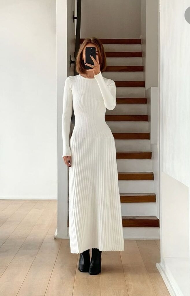 Person in a long white ribbed dress taking a mirror selfie on stairs, wearing black ankle boots.