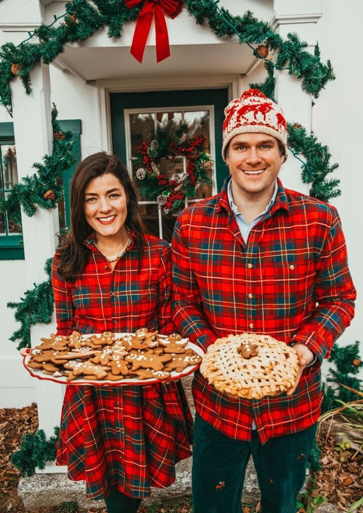 Smiling couple in red plaid outfits holding Christmas cookies and pie, standing by holiday wreath and garland.